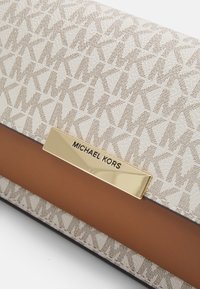 Beige clutch taske med monogrammønster, glat brun stribe og en guld logo-plade. Tekstureret overflade med elegante detaljer.