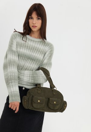 Ung kvinde i gråstribet sweater og sort nederdel, der holder en grøn stofhåndtaske med frontlommer, stående mod en ensartet hvid baggrund.