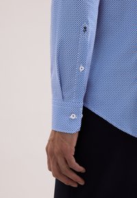 Niebieska koszula w geometryczny wzór, z białymi guzikami i prostym mankietem. Materiał ma gładką teksturę.