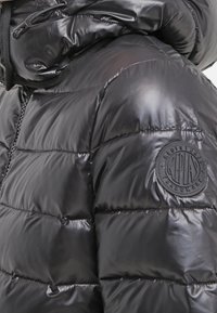 Black puffer jacket featuring a glossy finish, quilted design, and a circular logo patch on the sleeve. Textured, insulated material.