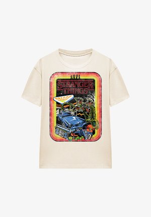 Beige cotton t-shirt featuring a colorful graphic print. Design includes a blue car, monsters, and items related to the "Stranger Things" theme.