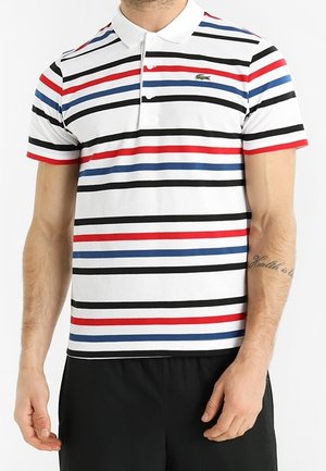 Uomo che indossa una polo bianca con strisce orizzontali rosse, nere e blu, e pantaloni neri, in piedi con le mani rilassate ai lati.