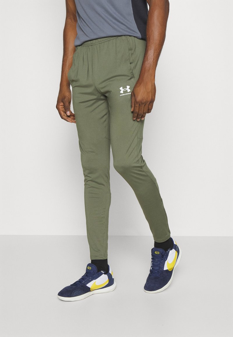 Zelene sportske hlače s elastičnim pojasom, sužene nogavice i bijeli Under Armour logo sprijeda, u kombinaciji s tamnim tenisicama.