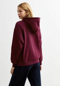 Felpa oversize bordeaux con cappuccio regolabile, polsini a costine e una texture morbida, mostrata dal retro. Abbinata a pantaloni scuri.