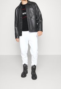 Chaqueta de cuero negra con bolsillos, camiseta gráfica negra, pantalones de chándal blancos y botas negras con cordones, con acentos plateados y logo.
