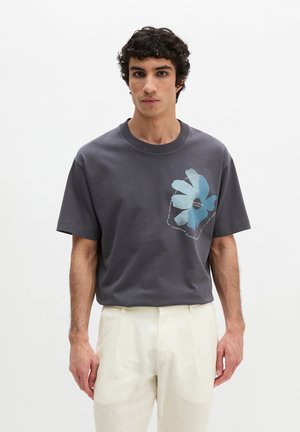 Jeune homme aux cheveux bouclés portant un t-shirt gris avec un motif de fleur bleue sur la poitrine et un pantalon beige clair, debout devant un fond blanc.