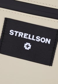 Beige tyg med en svart rektangulär etikett med texten "STRELLSON" i vit font. Etiketten har en vit schweizisk korslogotyp nedanför texten.