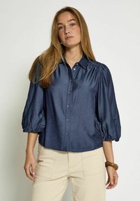 Chemise bleu marine à boutons avec des manches bouffantes et un col, fabriquée en tissu doux. Présente un ourlet droit et un éclat subtil.
