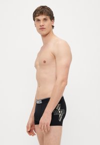 Melni bokseršorti no elastīga auduma ar baltu jostu, uz kuras ir uzraksts "DIESEL" un zelta grafiskais attēls sānos.