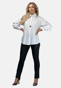 Biała koszula z guzikami z wysokim kołnierzem i dużymi guzikami z przodu, w zestawieniu z czarnymi dopasowanymi spodniami i wysokimi obcasami w wzór w kratę.