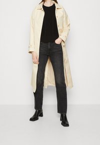Manteau trench couleur crème avec de grands boutons, associé à un pull texturé noir et à un jean noir. Des bottines noires à talons complètent la tenue.