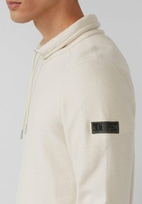 Kremowa bluza z kapturem o teksturowanej tkaninie, ze sznurkiem i czarną naszywką na lewym rękawie z napisem "Elevated Casual Est. 1969".