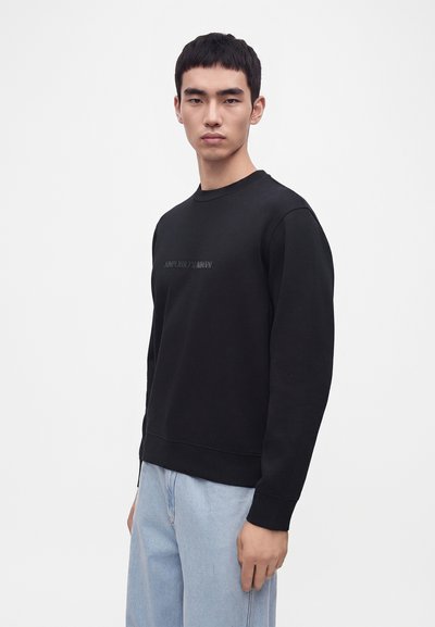 Jeune homme aux cheveux courts noirs portant un sweat-shirt noir à manches longues et un jean bleu clair sur un fond blanc uni.