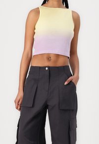 Vrouw in een mouwloze geel-tot-lila ombre crop top en hoge, donkere cargobroek met voor- en zijzakken.