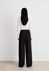 Pantalons larges noirs avec une texture lisse, assortis à une chemise blanche ajustée à manches longues. Vue de dos.