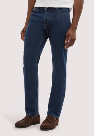 Mann trägt dunkelblaue Jeans mit geradem Bein, braune Loafers und ein weißes Hemd, steht vor einem schlichten hellen Hintergrund.