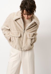 Beige faux fur jack met ritssluiting en een brede kraag, twee frontzakken en een zachte, textuurrijke oppervlakte. Draagt over een lichte trui en broek.