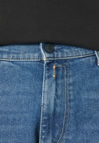 Μπλε τζιν παντελόνι με μαύρο κουμπί και αντίθετη ραφή. Ο λαιμός είναι ορατός στην κορυφή, με μια μικρή τσέπη με προσεγμένες άκρες.