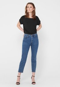 Svart kortermet topp kombinert med blå høytlivsjeans. Modellen har på seg svarte hæl-sandaler med et enkelt design og naturlig hår.