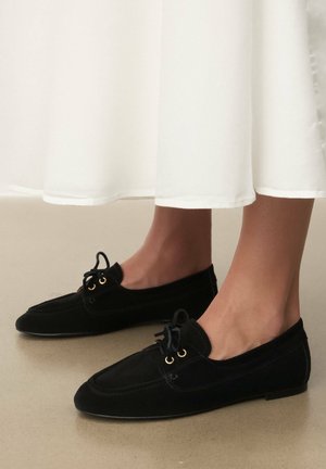 Črni semišasti mokasini s koničastim prstom, z zlatimi očesi in črnimi vezalkami. Čevlji so postavljeni na svetlo podlago.