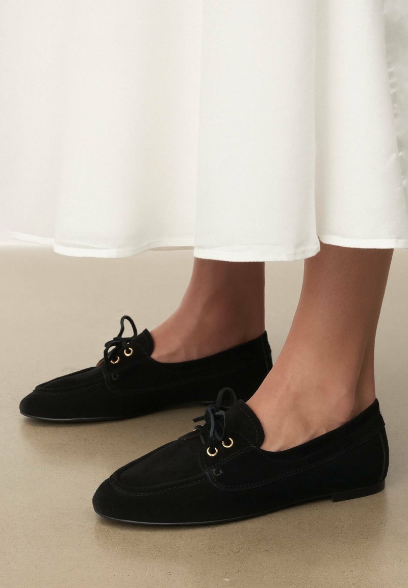 Mocassins en daim noir avec un bout pointu, dotés d'œillets dorés et de lacets noirs. Les chaussures sont posées sur une surface de couleur claire.