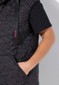 Chaleco acolchado negro con costuras rosas y patrones de estrellas, que cuenta con un bolsillo delantero y capucha. Llevado sobre una camiseta negra de mangas cortas.