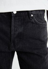 Pantalones vaqueros de mezclilla negra con un diseño clásico de cinco bolsillos, costuras visibles y herrajes de botón plateado en la cintura. Textura suave.