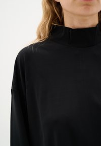 Čierny top s vysokým golierom vyrobený z hladkého, ľahkého materiálu; má voľný strih a spadnuté ramená, čo ponúka minimalistický dizajn.