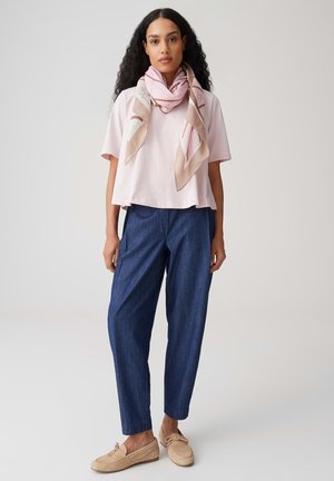 Woman with long curly hair wearing a light pink blouse, patterned scarf, dark blue pants, and beige loafers standing against a plain background.