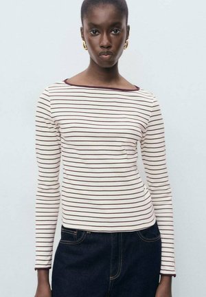Young woman wearing gold hoop earrings, a white long-sleeve shirt with thin horizontal maroon stripes, and dark blue jeans.