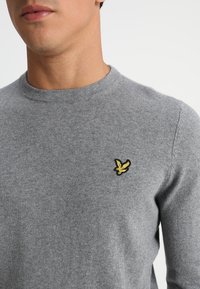 Maglione grigio realizzato in morbido materiale, con collo rotondo e un piccolo logo di un uccellino giallo ricamato sul petto.