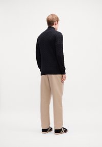 Fekete kötött pulóver magas gallérral, bézs nadrággal párosítva, hátulról nézve. A cipők feketék fehér részletekkel.