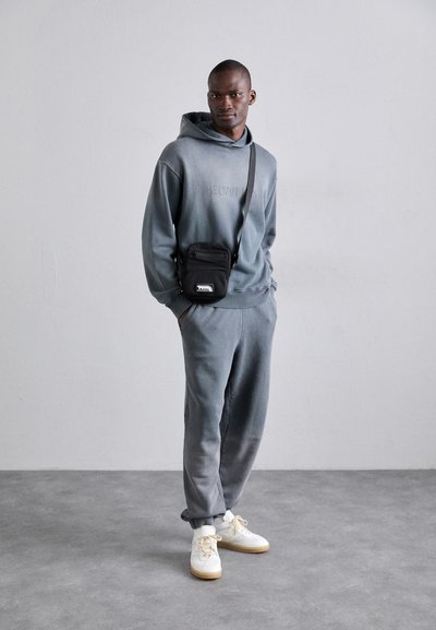 Sweatshirt à capuche gris avec un léger dégradé, associé à un pantalon de survêtement assorti et des baskets blanches. Un petit sac crossbody noir est porté.