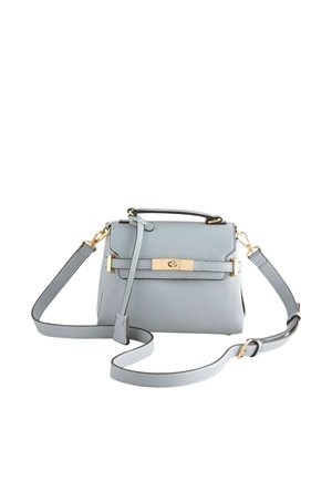 Junge Frau mit dunklen Haaren trägt eine schwarze Lederjacke, ein schwarzes Top, goldene Ohrringe und ein Armband und hält eine hellgraue Handtasche mit goldenen Beschlägen.