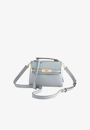 Jeune femme aux cheveux foncés porte une veste en cuir noire, un haut noir, des boucles d'oreilles et un bracelet dorés, tenant un sac à main gris clair avec des accessoires dorés.