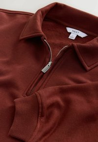 Hnědý svetr s čtvrtím zipem a límcem, stříbrný zip částečně rozepnutý, žebrované náplety a štítek značky "NEXT" uvnitř límce.