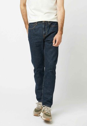 Mann trägt dunkelblaue Jeans mit geradem Bein, weißes T-Shirt und beigefarbene Sneakers, steht vor weißem Hintergrund.