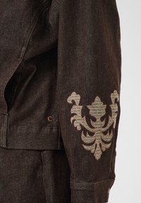 Manche de veste en denim brun foncé avec un motif brodé floral beige orné près du coude et des détails de couture visibles.