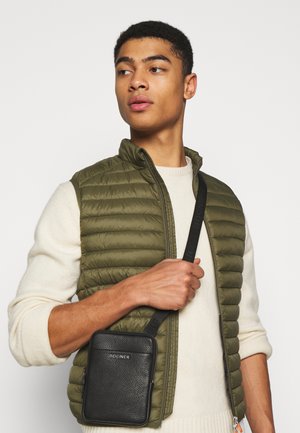 Black leather crossbody bag with a textured surface and zippered closure, worn with an olive quilted vest over a cream sweater.