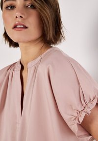 Lockere Bluse in Blassrosa mit rundem Ausschnitt, kurzen Puffärmeln und dezenten Raffungen. Glatte Stoffoberfläche und klare Linien.