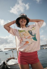 Svetlo roza prevelika majica s pisanim grafičnim dizajnom na sprednji strani, združena z rdečimi kratkimi hlačami s črtami. Priložnostni slog, primeren za toplo vreme.