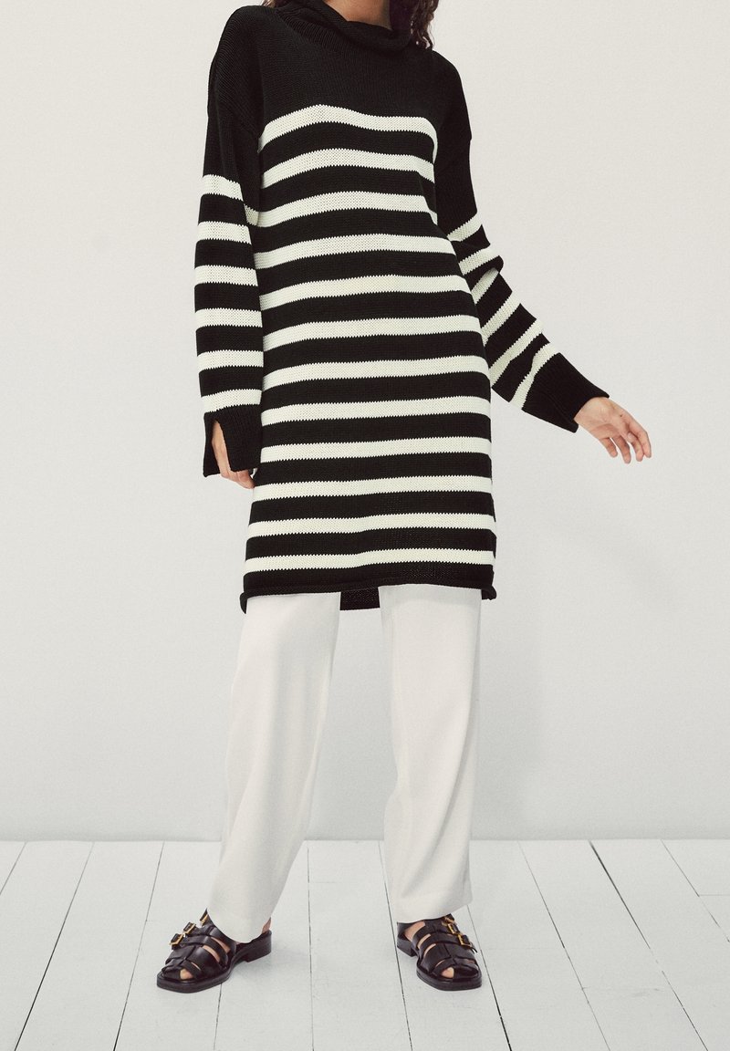 Personne portant une robe pull rayée horizontale noire et blanche, un pantalon blanc ample et des sandales noires avec des boucles, debout sur un parquet en bois blanc.