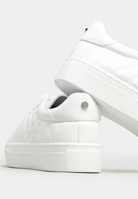 Zapatillas blancas acolchadas con suela de goma texturizada. Presentan una puntera redondeada, diseño de cordones y un acento metálico en el talón.