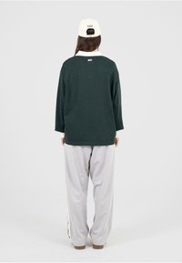Sudadera oversized verde oscuro con mangas largas, textura suave y puños blancos; combinada con pantalones gris claro. Diseño sencillo, sin logotipos visibles.