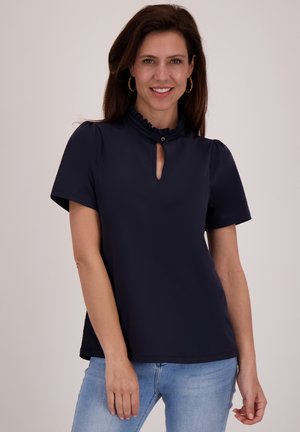 Donkerblauw shirt met korte mouwen en een gerimpelde opstaande kraag, met een keyhole-opening en gladde stof. Gecombineerd met lichte spijkerjeans.