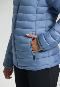 Personne portant une veste matelassée bleu clair avec une poche zippée, la main placée à l'intérieur de la poche, associée à un pantalon noir.