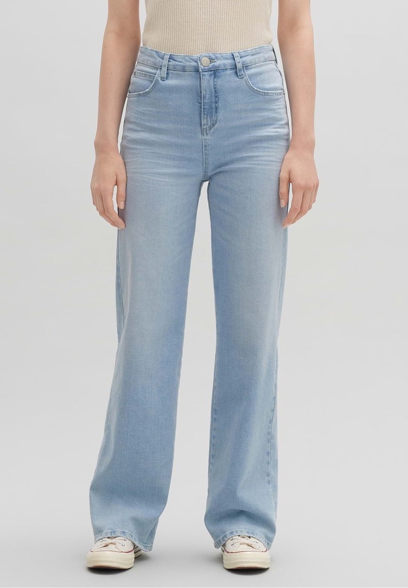 Persona vestida con jeans de tiro alto y piernas rectas de color azul claro, con zapatillas blancas y una camiseta sin mangas beige, de pie frente a un fondo liso.