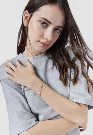 Pulsera de plata con charms de estrella y flor, llevada en un brazo de piel clara. Camiseta gris de gran tamaño con mangas dobladas en el fondo.