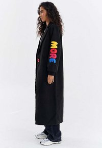 Kobieta stojąca bokiem, ubrana w długi czarny płaszcz z kolorowymi literami "MORE" na rękawie, ciemne spodnie i białe trampki.