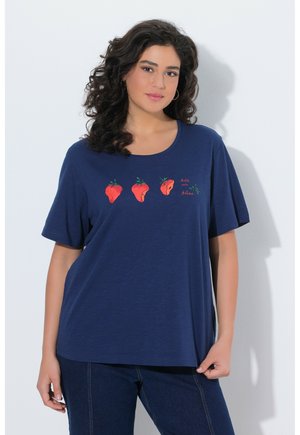 Kvinde med krøllet mørkt hår iført marineblå T-shirt med tre rådne jordbær og teksten "bite into bliss", kombineret med mørke jeans.